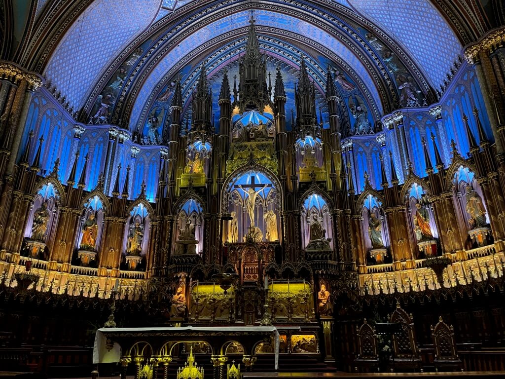 The chancel (front of the sanctuary) of Notre Dame in Montreal, Quebec, Canada. The crucifix and other statues are lit with colorful lights, many of them in blue hues.