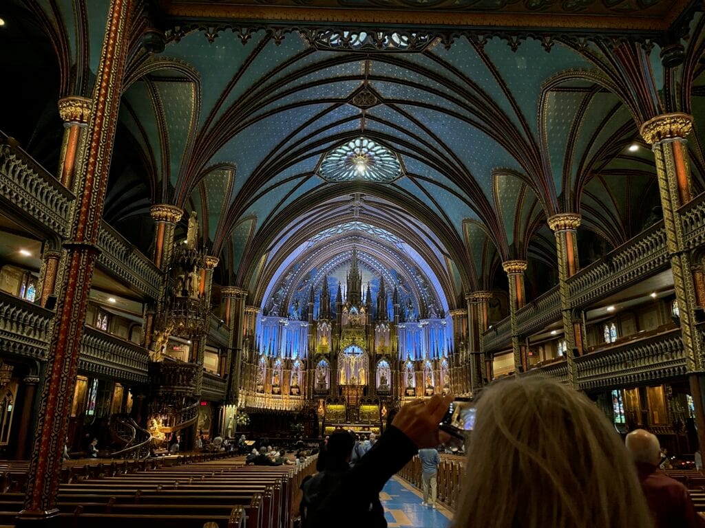 The sanctuary of Notre Dame cathedral in Montreal, Quebec, Canada. It's a gigantic, vaulted ceiling with much ornate artwork. The interior is lit with colored lighting. In the foreground, another photographer is taking a photo on their phone.