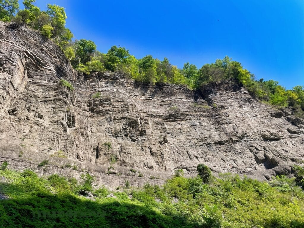 A gray rock cliff towers above with green vegetation at the bottom and top. The sky is all blue. We can almost hear the voices of the earth when we see sights like this.