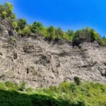 A gray rock cliff towers above with green vegetation at the bottom and top. The sky is all blue. We can almost hear the voices of the earth when we see sights like this.