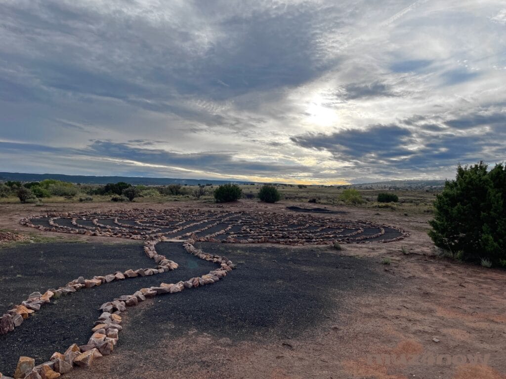 The sun barely cuts through heavy cloud cover. On the red desert sands below, there is a spiral trail. It would be difficult to go off the path in this spiral since large stones run along the edge of it.