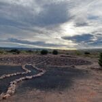 The sun barely cuts through heavy cloud cover. On the red desert sands below, there is a spiral trail. It would be difficult to go off the path in this spiral since large stones run along the edge of it.