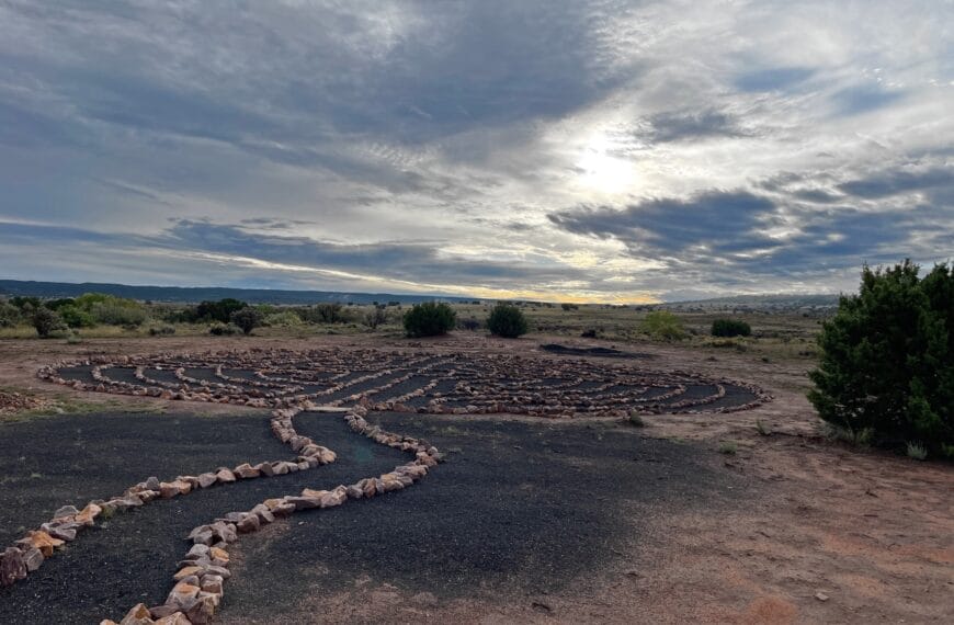 The sun barely cuts through heavy cloud cover. On the red desert sands below, there is a spiral trail. It would be difficult to go off the path in this spiral since large stones run along the edge of it.